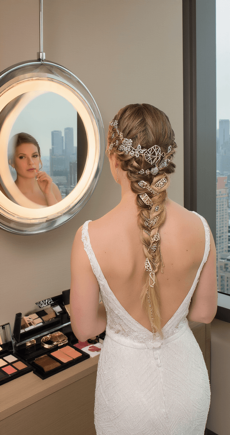 A bride in a modern column dress inspects her Dutch crown braid adorned with mixed metallic hair accessories in a contemporary hotel suite, with city skyline views visible through a suspended circular mirror; a makeup station with luxury products is in the background.