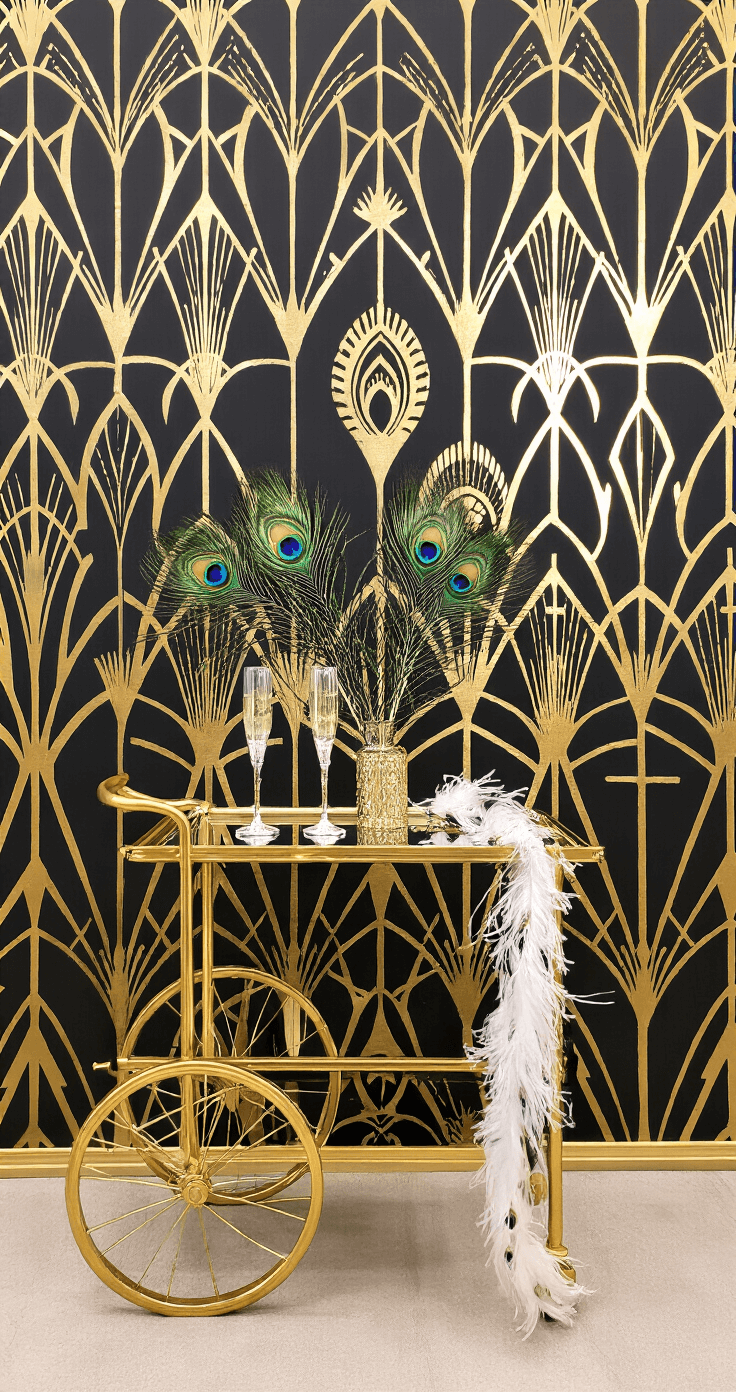 An Art Deco glamour station featuring a black and gold geometric backdrop with peacock feather accents, a mirror-topped gold bar cart with champagne glass props and feather boas, and dramatic uplighting casting geometric shadows, all shot from above to highlight the floor pattern reflection in rich jewel tones and metallic accents.