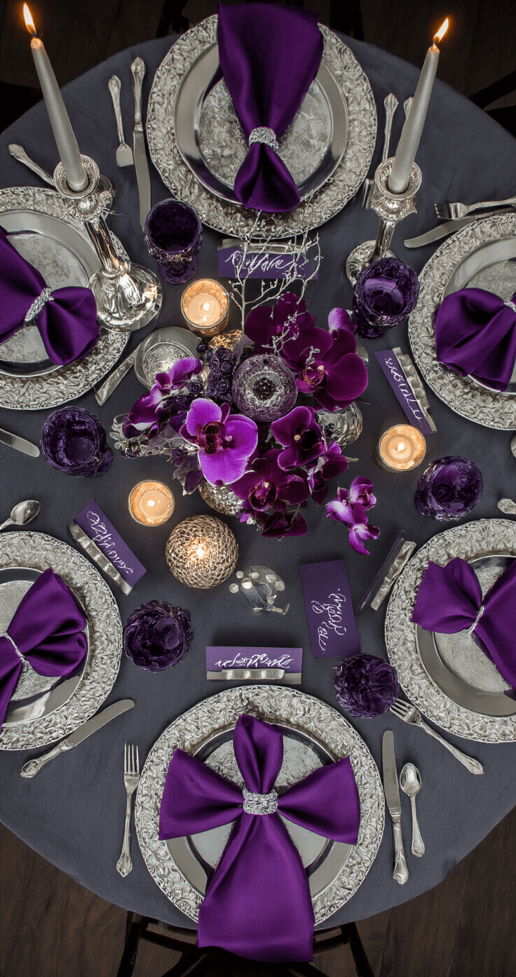 The Ultimate Guide to November Wedding Colors That Will Make Your Special Day Absolutely Stunning Dramatic overhead view of a November wedding tablescape featuring amethyst purple and silver accents, including a round table for eight with moody lighting, charcoal grey linen, layered silk napkins, a centerpiece of deep purple orchids, and glowing silver candelabras against a dark wooden floor.